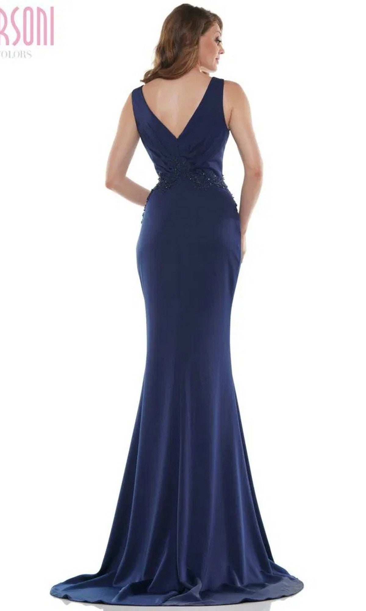 A woman stands with her back to the camera, showcasing the Marsoni MV1054 Long Formal Dress: a sleeveless, floor-length navy gown with a V-shaped back, subtle waist embellishments, and an elegant trumpet silhouette.